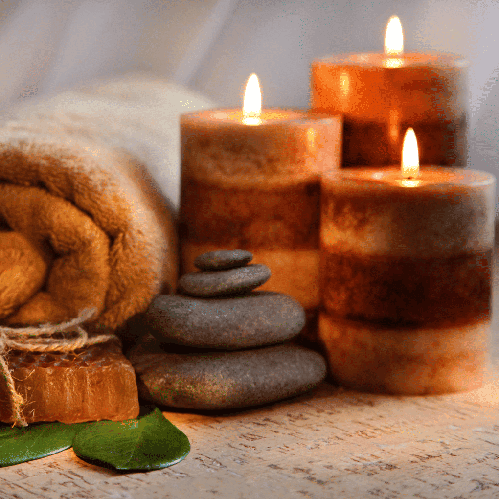 Spa setting with lit candles, stacked stones, rolled towel, soap, and green leaf.