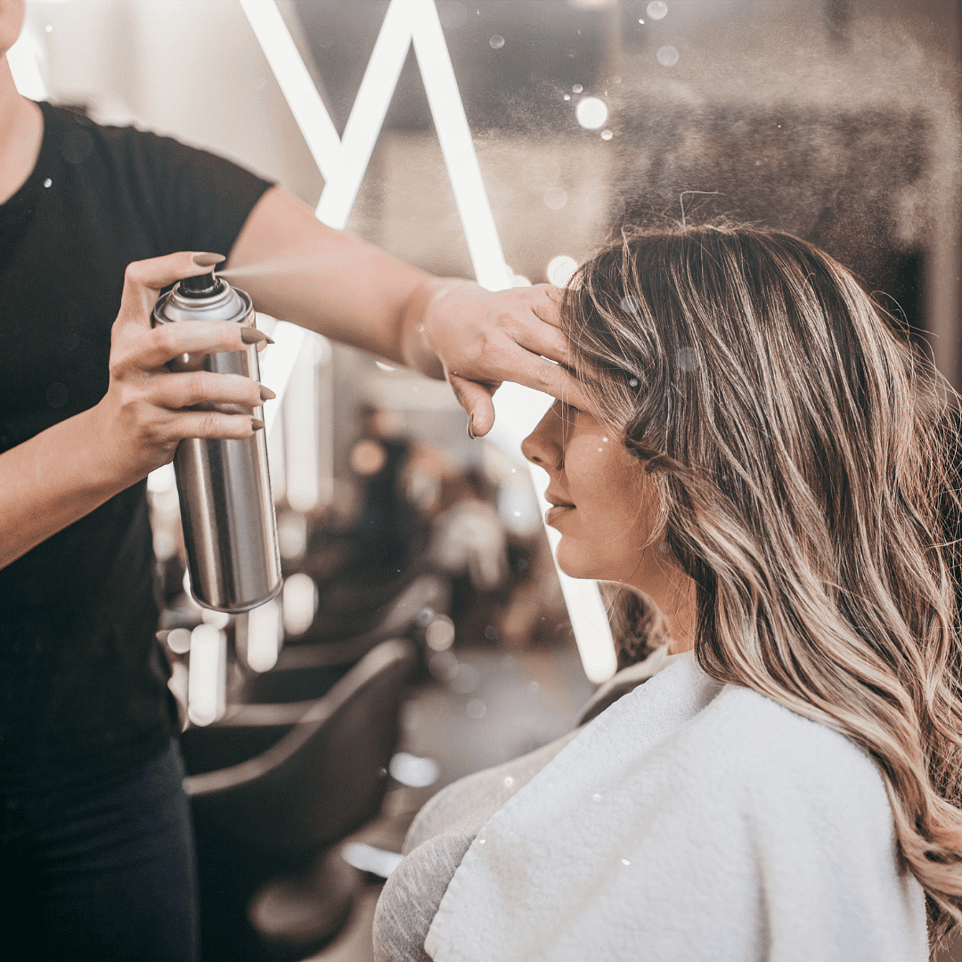 Stylist applying hairspray to a woman's hair in a salon.