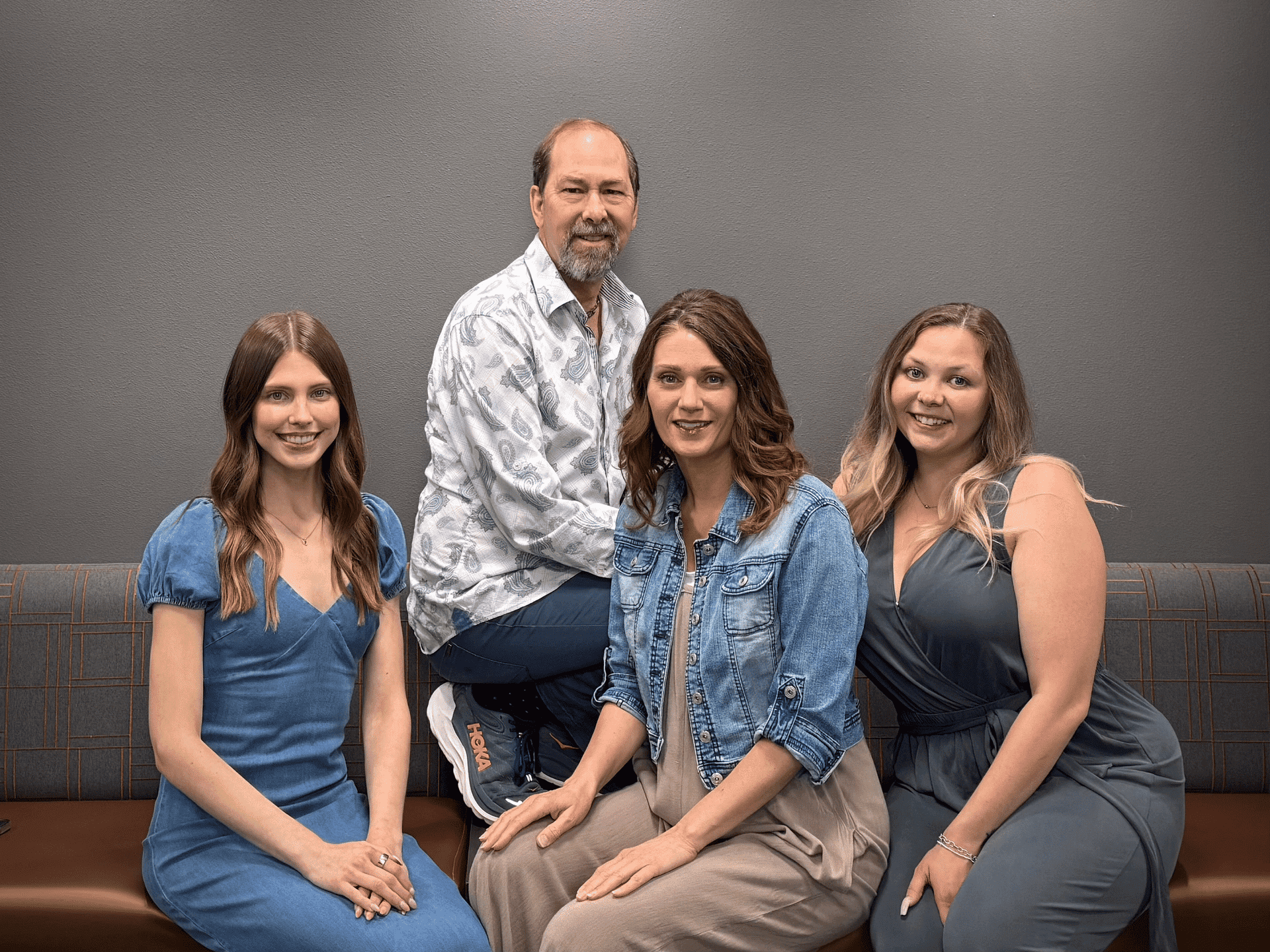 Four adults posing together, sitting on a bench against a gray wall.