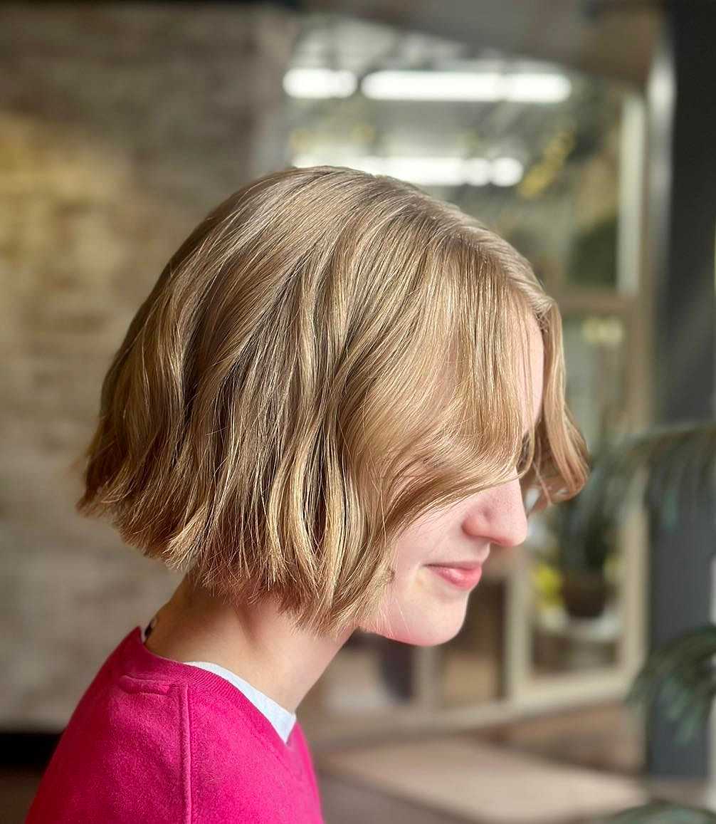 Profile of a person with short, wavy blonde hair, wearing a pink shirt, against a blurry background.